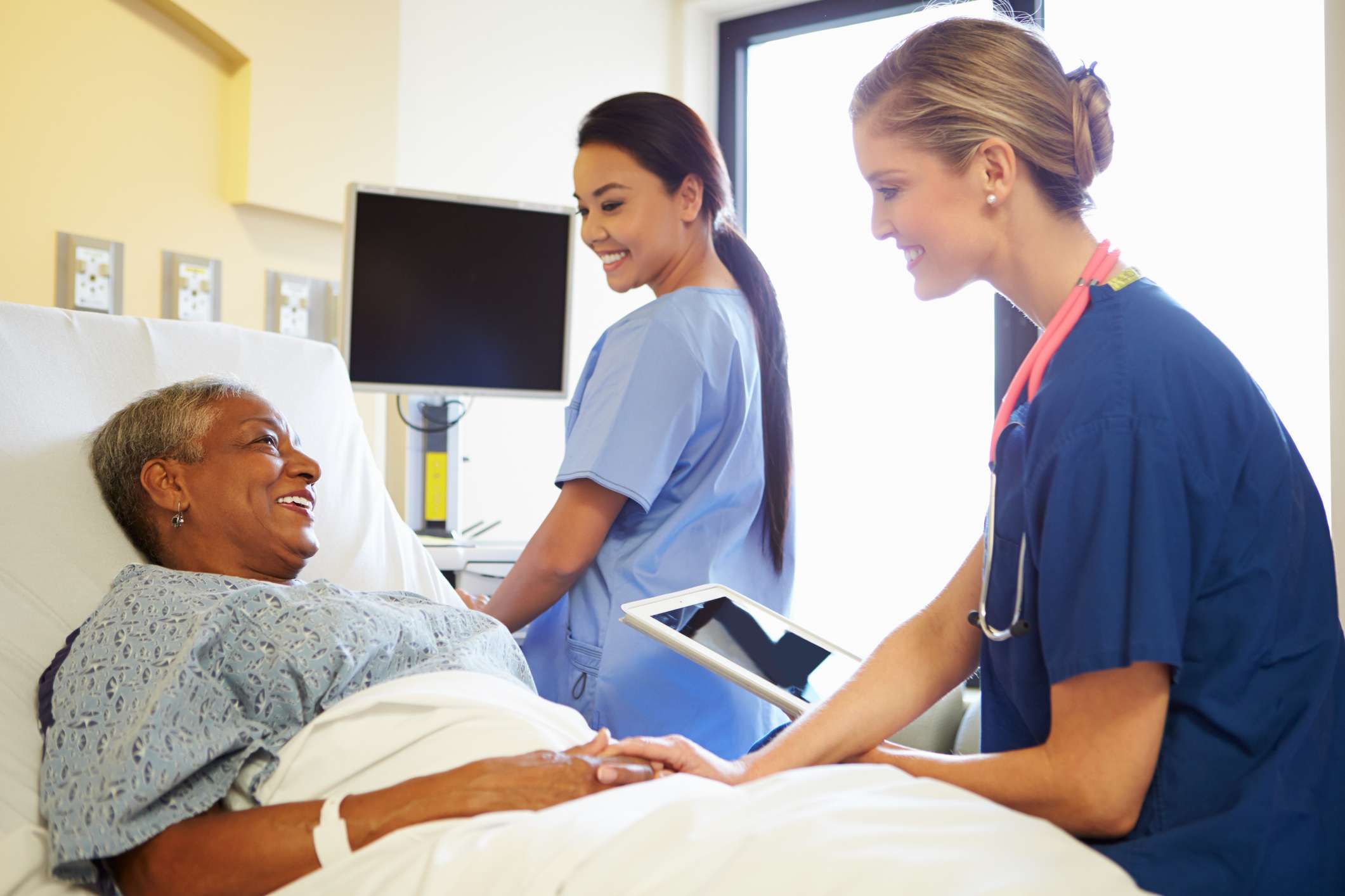 Smiling Nurse With Digital Tablet Talks To Female Patient In Hospital Bed